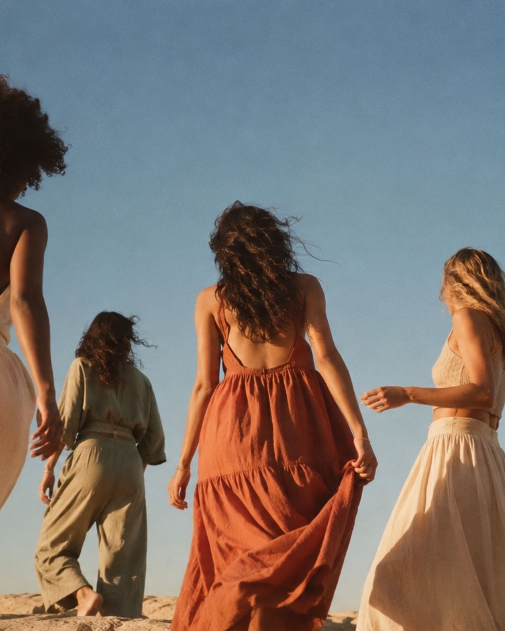 Quatre femmes marchant ensemble face à un ciel bleu, robes fluides au vent
